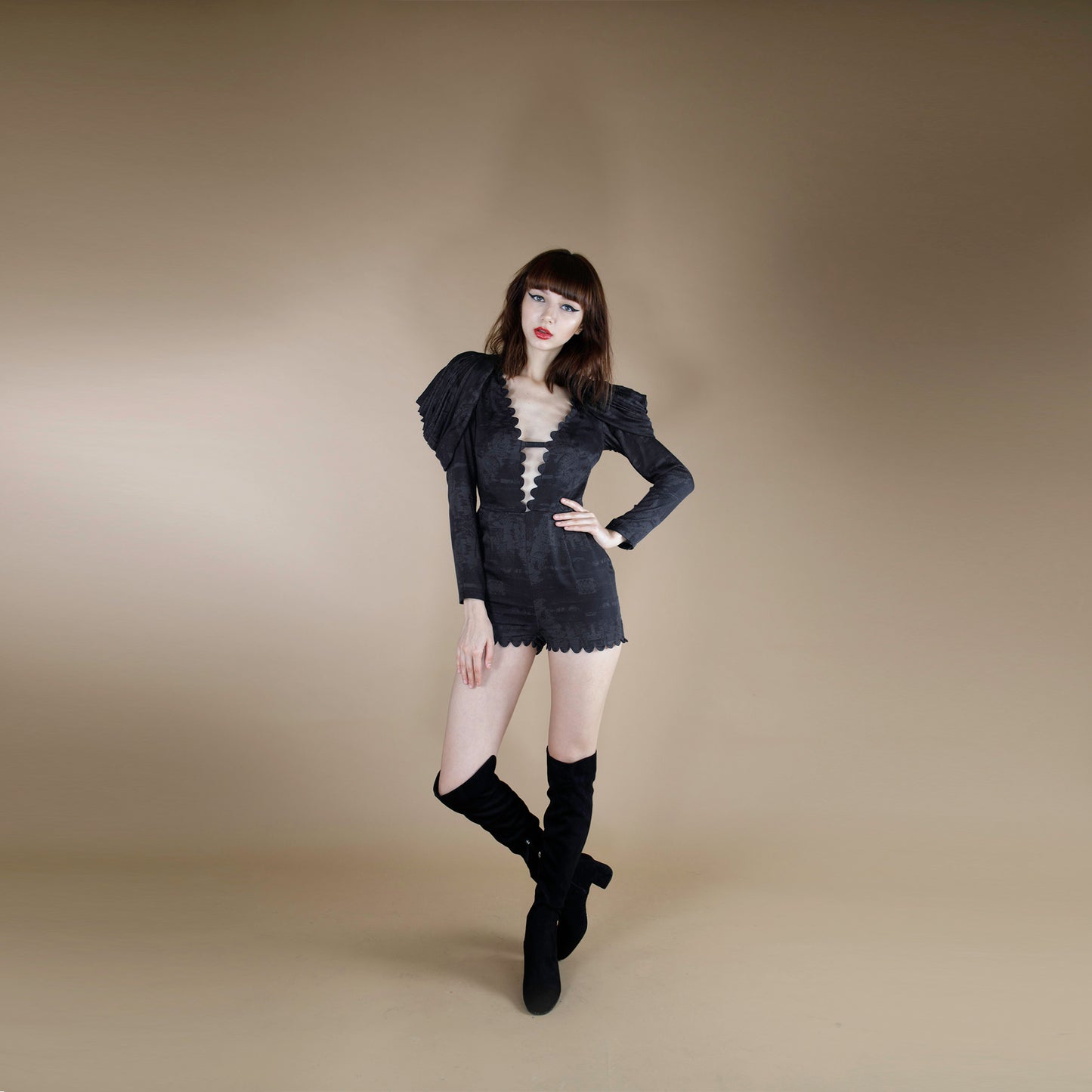 A global small size model wearing a black tonal print short jumpsuit accentuated with cowl cape sleeves. the jumpsuit is made from organic lotus stem silk fabric and adorned with a scallop neck line and hem.  