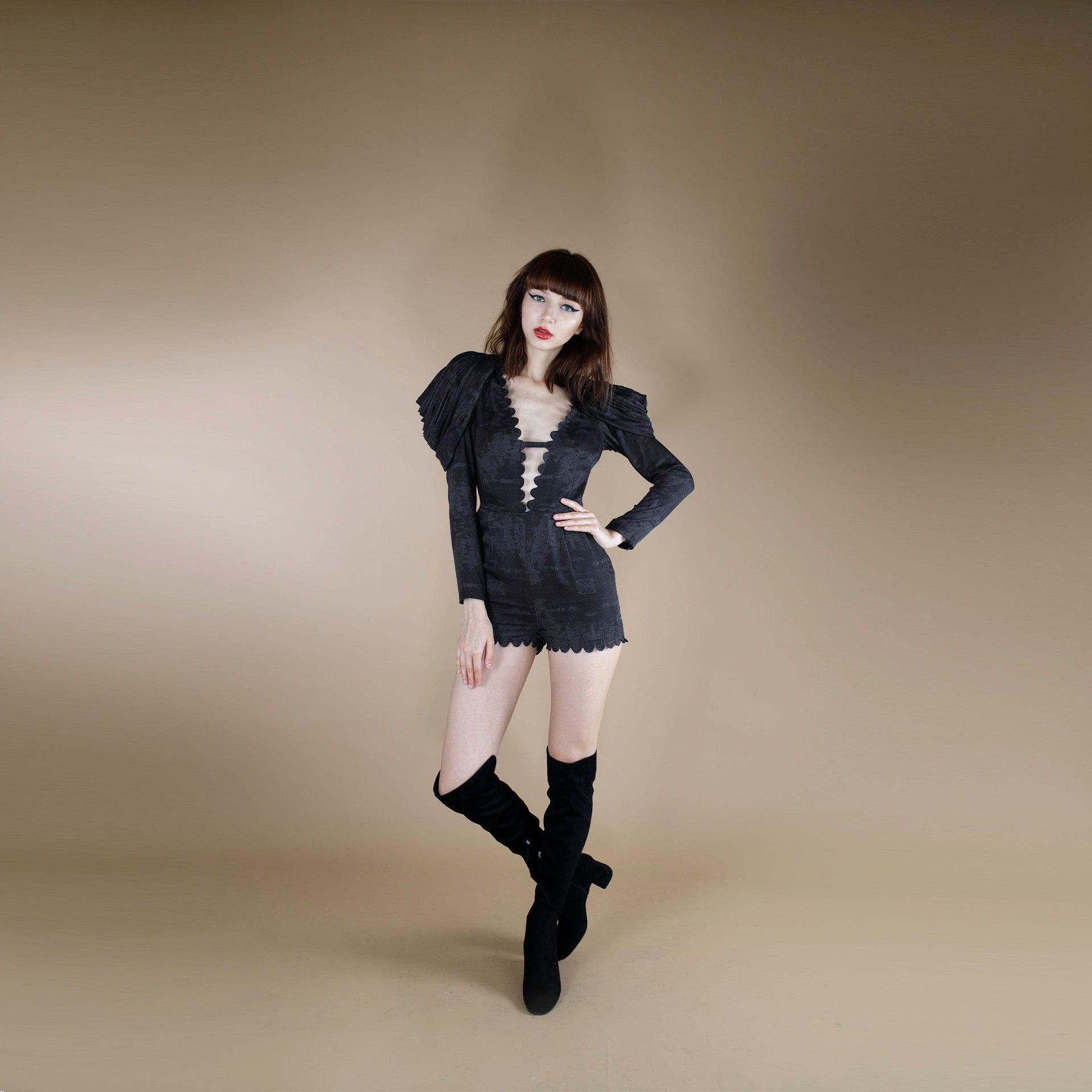 A global small size model wearing a black tonal print short jumpsuit accentuated with cowl cape sleeves. the jumpsuit is made from organic lotus stem silk fabric and adorned with a scallop neck line and hem.  