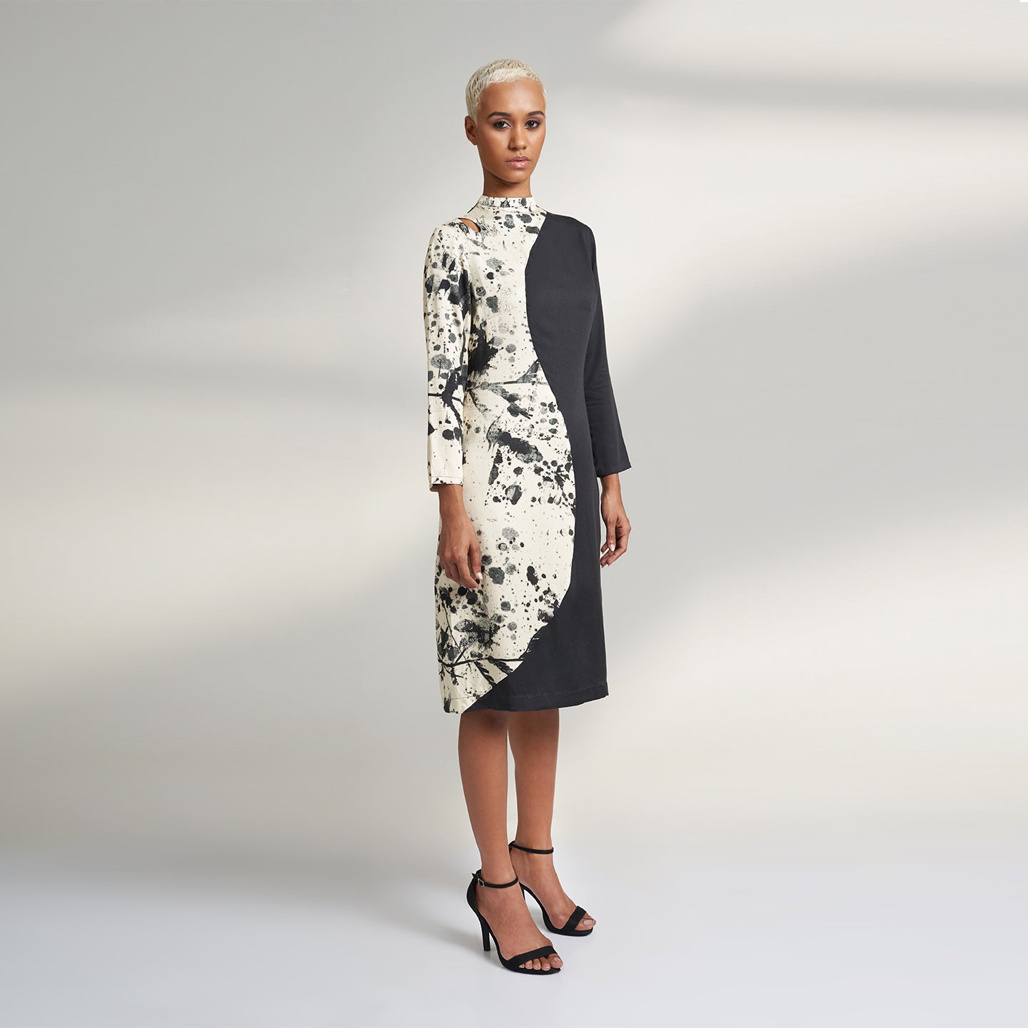 A GLOBAL SMALL SIZE MODEL wearing a black and cream midi dress with one side in solid black and other side in cream with black print on it. the dress is highlighted with a key hole on the right shoulder. 