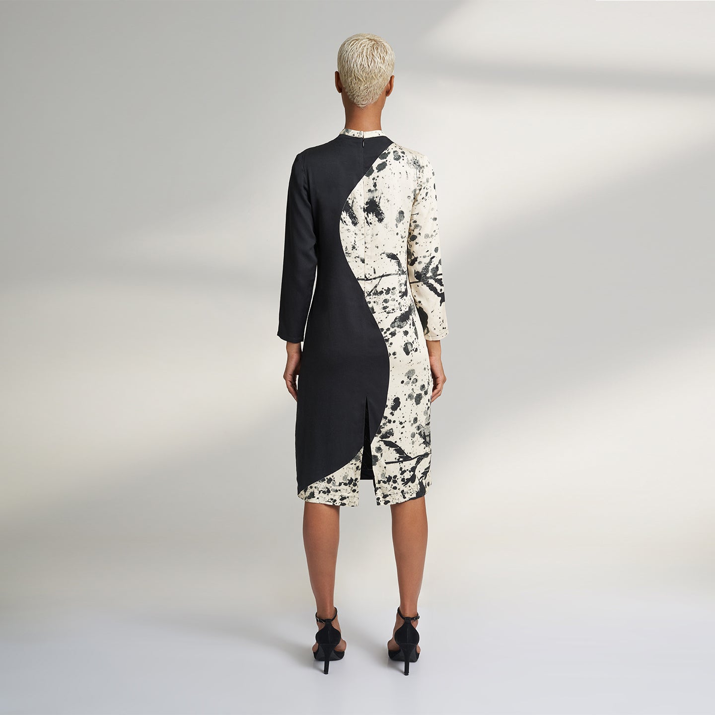 A GLOBAL SMALL SIZE MODEL wearing a black and cream midi dress with one side in solid black and other side in cream with black print on it. the dress is highlighted with a key hole on the right shoulder. 